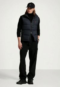 Mies, który ma na sobie czapkę w kolorze czarnym, czarny golf, granatową kamizelkę puchową, czarne spodnie i czarne sneakersy, stoi z rękami w kieszeniach.