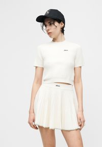 Νεαρό άτομο που φορά λευκό ριπ ζιβάγκο τοπ και πλισέ φούστα με λογότυπα επωνυμίας, συνδυασμένο με μαύρο καπέλο μπέιζμπολ, στέκεται μπροστά σε έναν απλό φόντο.