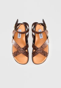 Sandalias marrones con estampado y tiras cruzadas, hechas de cuero texturizado. Hebilla ajustable en el tobillo y plantilla acolchada. Suela plana.