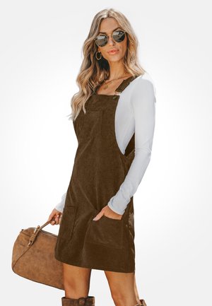 Femme aux longs cheveux blonds portant des lunettes de soleil rondes, une chemise blanche à manches longues, une robe salopette en velours côtelé marron, tenant un sac à main marron.