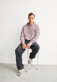 Uomo con i capelli corti e la barba seduto su uno sgabello di metallo, indossando una felpa lavanda, jeans neri larghi e sneakers grigie, su uno sfondo bianco uniforme.