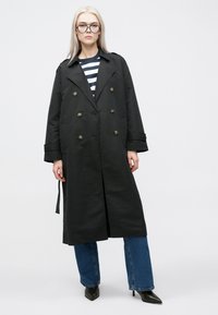 Zwarte dubbelgeknoopte trenchcoat met grote knopen, zijzakken en een ceintuur in de taille, gecombineerd met een gestreept shirt en wijde jeans.