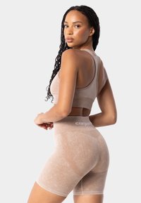 Béžová, přiléhavá sportovní podprsenka a kratasy s vysokým pasem, vyrobené z texturované tkaniny. Obsahuje žebrovaný pás a logo značky na pasu, bezešvý design.