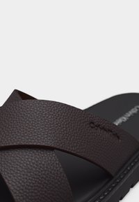 Καφέ δερμάτινο σανδάλι με σταυρωτούς ιμάντες και υφή στην επιφάνεια, με μαύρη σόλα και σήμανση Calvin Klein στον επάνω ιμάντα.