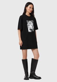 Vestido camiseta negro oversize con estampado gráfico de un león y el texto "ALLSAINT'S", combinado con botas de cuero negras hasta la rodilla.