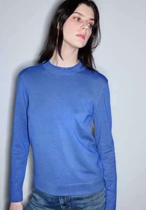 Jonge vrouw met donker haar, gekleed in een blauw truitje met lange mouwen en een spijkerbroek, die iets naar de zijkant kijkt tegen een effen achtergrond.