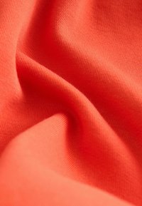 Záber zblízka na mäkkú, hladkú oranžovú látku s viditeľnou jemnou pletenou textúrou a jemnými záhybmi vytvárajúcimi subtílne tiene a odlesky.