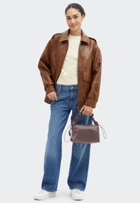 Brown leather jacket, light yellow shirt, blue jeans, and white sneakers. Holding a small, dark gray bag with twisted handles.