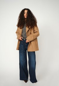 Tan wool double-breasted coat, grey t-shirt beneath, blue wide-leg denim jeans, and pointed brown heels. Soft texture and clean lines.