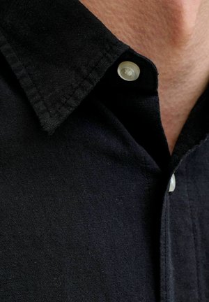 Pritlični posnetek ovratnika črne srajce z dvema belima gumboma na svetli koži.