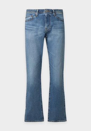 Ung mand iført en beige T-shirt med korte ærmer, blå jeans og brune loafers stående mod en enkel lys baggrund.