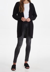 Černý pletený kardigan s otevřeným předním dílem, texturovaný vzor, v kombinaci s bílou košilí a tmavě šedými skinny džíny. Černé klouzavé sandály.