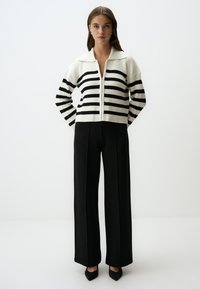 Cardigan a righe bianche e nere in maglia con colletto, abbinato a pantaloni a vita alta neri larghi e tacchi a punta neri.
