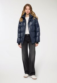 Giacca imbottita blu navy con finitura lucida, dotata di colletto alto, bottoni a pressione e polsini elasticizzati, abbinata a jeans neri a gamba larga e sneakers bianche.