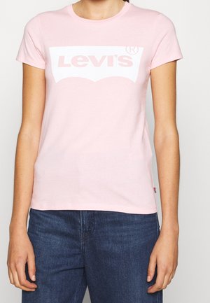 Osoba ubrana w jasnoróżową koszulkę Levi's z białym logo i ciemnoniebieskie dżinsy, widoczna od szyi do bioder na jednolitym tle.