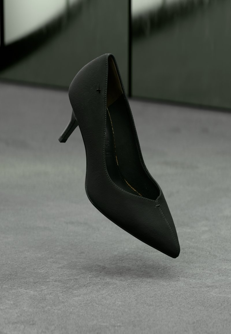 Μαύρο ψηλοτάκουνο παπούτσι με μυτερή μύτη να αιωρείται πάνω σε γκρι χαλιού με φόντο έναν σκοτεινό πράσινο τοίχο.