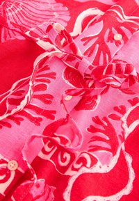 Klarröd blus med blommönster i olika nyanser av rosa. Har en knytdetalj med rosett och små knappar längs fronten. Linne textur.