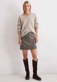 Beżowy sweter dzianinowy z dekoltem w serek, mini spódnica w kratę o fakturowanej tkaninie w odcieniach brązu i czerni oraz wysokie brązowe zamszowe kozaki na niewielkim obcasie.