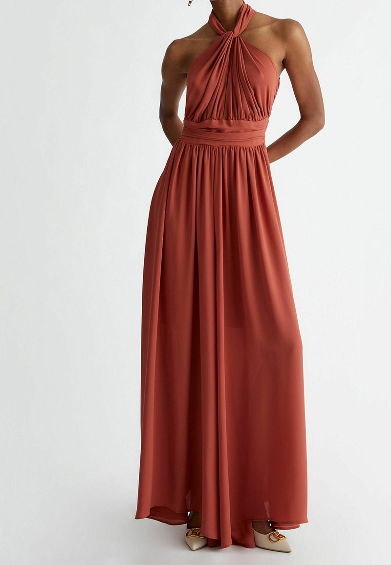 Femme portant une robe longue couleur rouille avec un col licou, un corsage froncé et une large ceinture, associée à des chaussures blanches pointues.