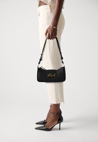 Black leather handbag with gold logo, rectangular shape, short handle, worn with ivory cropped jeans and black pointed-toe heels.