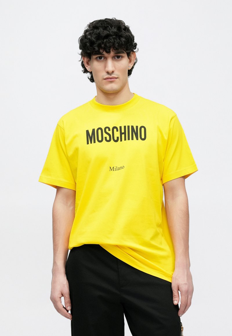 Gul bomuld T-shirt med fed sort "MOSCHINO" tekst og mindre "Milano" tryk, afslappet pasform, korte ærmer og rund halsudskæring.