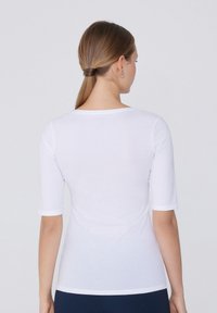 Camicia bianca aderente con maniche a tre quarti, caratterizzata da un collo rotondo e una texture liscia e morbida. Nessun motivo o accentuazione visibili.