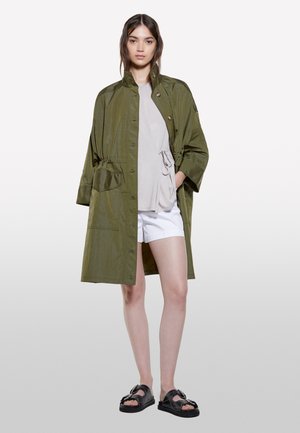 Jeune femme portant un manteau boutonné vert olive sur un haut gris clair et un short blanc, debout, les mains dans les poches et des sandales noires.