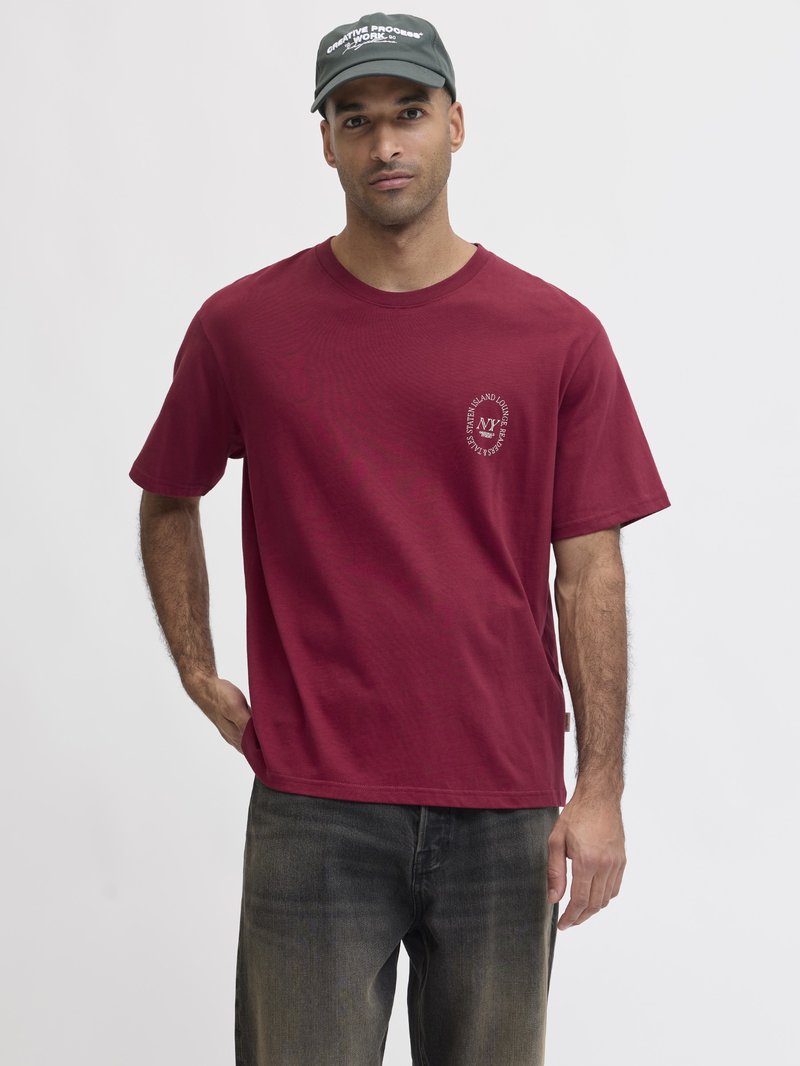 Man wearing a burgundy T-shirt with circular white text on the left chest and a dark green cap with white text, standing against a plain background.