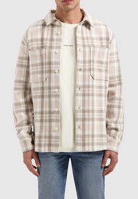 Beige en bruine geruite shirt met een knoopsluiting aan de voorkant, twee borstzakken en een zachte textuur, gedragen over een lichte t-shirt.