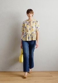 Floral Peplum-Top in Gelb mit blauen Akzenten, kombiniert mit ausgestellten blauen Jeans. Das Model hält eine gelbe Handtasche und steht auf einem Holzfußboden.