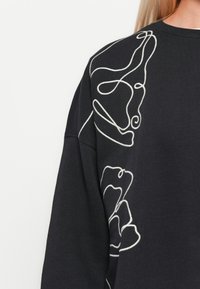 Svart hettegenser med hvite sammenhengende linjeabstrakte ansiktsdesign på skulderen og overarmen.