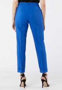 Pantalon bleu sur mesure à coupe ajustée, ourlets retroussés, avec deux poches arrière, associé à des sandales à talons noirs. Texture lisse.