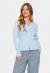 Světle modrý cardigan s výstřihem do V, ribbovanou strukturou a sedmi knoflíky vpředu. Obsahuje volánkový lem a přiléhavé dlouhé rukávy.