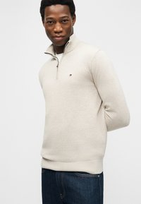 Νεαρός άνδρας με σκούρο δέρμα, ντυμένος με μπεζ πουλόβερ με φερμουάρ μέχρι το στήθος και σκουρό μπλε τζιν, όρθιος με τα χέρια πίσω από την πλάτη.