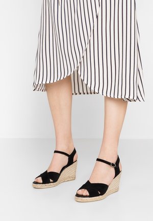 Czarne sandały na koturnie zamszowe z podeszwą z juty, otwartym noskiem i regulowanym paskiem wokół kostki. Stylizowane z paskowaną spódnicą.