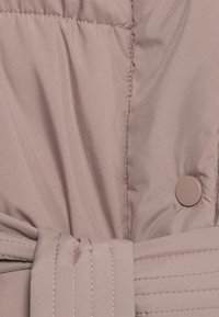 Närbild av beige quiltad tyg med sydd söm, en rund knapp och ett matchande tygbälte knutet i en knut.