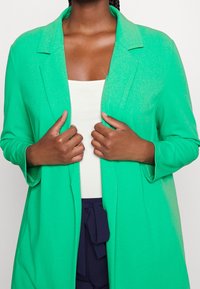 Zielona marynarka o gładkiej fakturze, z długimi rękawami i wyciętym kołnierzem. Nosi się ją na jasnym topie na ramiączkach, w połączeniu z ciemnymi spodniami.