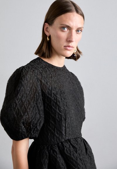 Black textured dress with puffed sleeves, high neckline, and gathered waist. Sheen adds depth to the fabric's wavy pattern. Gold and purple earrings.