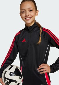 Schwarze Sportjacke mit roten Streifen, hohem Kragen und weißen Akzenten. Hält einen schwarz-weißen Fußball, geflochtenes Haar sichtbar.