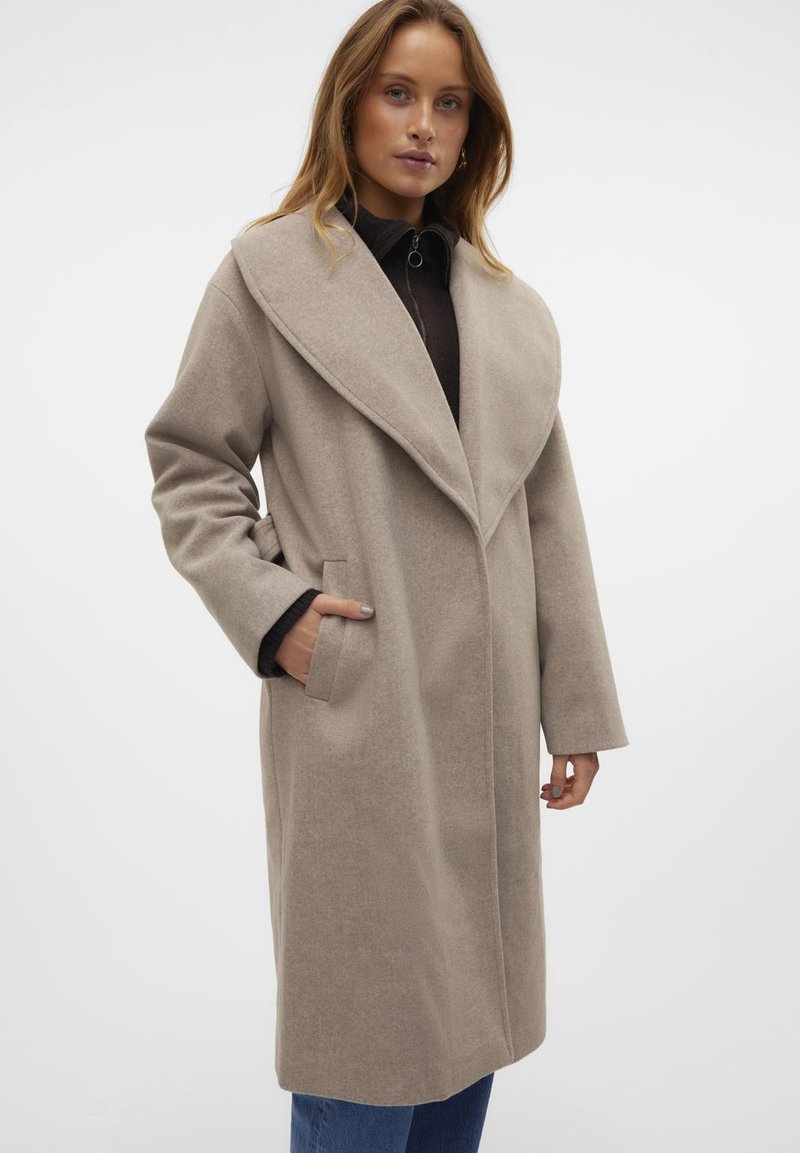 Cappotto in misto lana beige con ampio colletto, maniche lunghe, tasche laterali e vestibilità comoda, indossato sopra un top scuro e jeans blu.