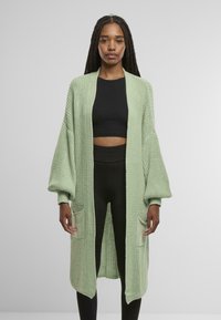 Ljusgrön stickad cardigan med långa ballongsärmar, öppen framtill och två framfickor, bärs över en svart crop top och leggings.
