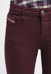 Närbild av en person som bär vinröda jeans med silvrig knapp och rutigt skjorta instoppad.