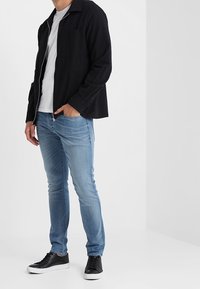 Chaqueta negra con cremallera sobre una camiseta blanca, jeans ajustados azules y zapatillas negras. Enfóquese en el estilo casual en capas y la textura del denim.