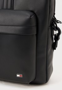 Black backpack made of smooth and textured materials, featuring a ribbed bottom panel and a small logo accent on the side.