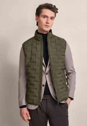 Grüne, gesteppte Pufferweste mit hohem Kragen, getragen über einem grauen Blazer und einem schwarzen Rollkragenpullover. Die Knöpfe am Blazer setzen einen detaillierten Akzent.