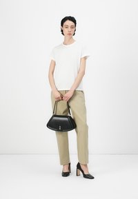 Schwarze Handtasche mit einer strukturierten, dreieckigen Form, die über ein goldfarbenes Hardware-Detail verfügt. Das Modell trägt ein weißes T-Shirt und beige Hosen.