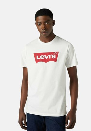 Joven con una camiseta blanca de Levi's con un logo rojo, de pie contra un fondo claro y uniforme, mirando al frente con una expresión neutral.