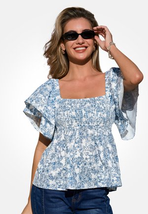 Lächelnde Frau mit schwarzen Sonnenbrillen, blau-weiß geblümter Bluse mit quadratischem Ausschnitt und flatternden Ärmeln sowie dunkelblauer Jeans, die ihre Sonnenbrille berührt.
