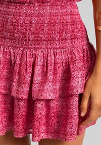 Jupe en tissu rose avec un smocking texturé à la taille, des volants superposés et un motif floral dans des nuances variées de rose et de rouge.