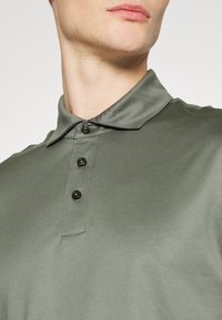 Grön polotröja med en jämn textur, som har en vikbar krage, tre svarta knappar och en figurnära design, vilket visar halslinning och övre tyg.